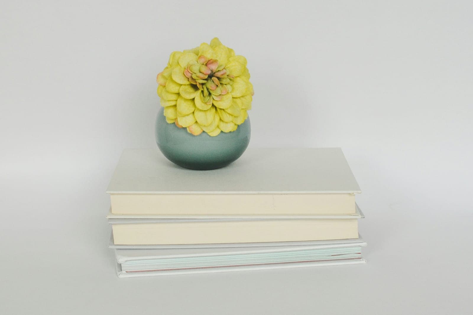 A small sage-green bud vase with a yellow flower on a stack of three plain cream hardcovers.