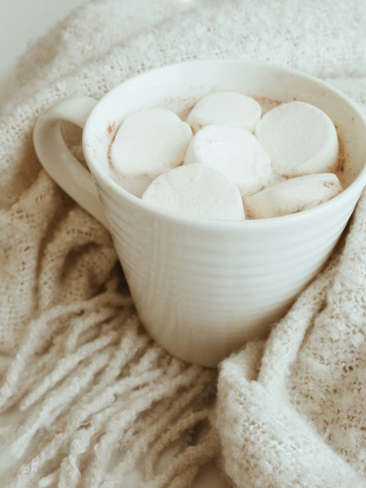 A white mug of hot cocoa topped with large marshmallows, in a cream knit blanket.
