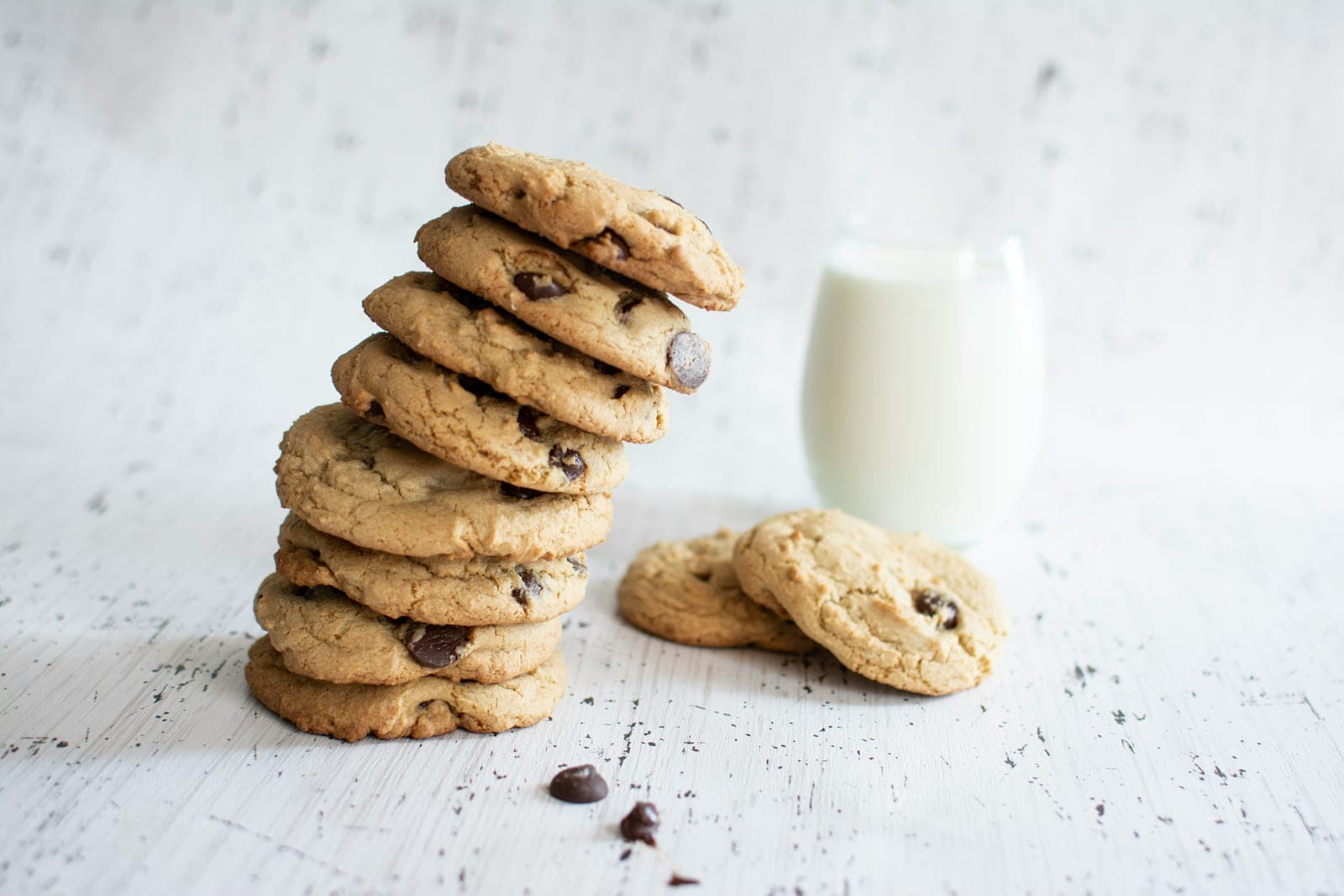 A stack of chocolate chip cookies beside a glass of milk on a white wooden table.