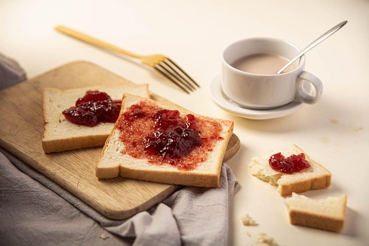 Slices of soft white bread spread with strawberry jam on a light wooden board, a cup of tea behind them.