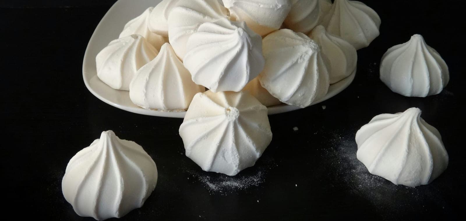 White piped meringues piled on a plate and scattered on a dark table.