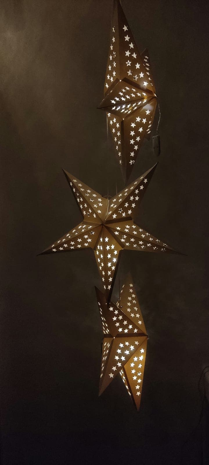 Three gold paper star lanterns pierced with tiny stars, hanging one above another against a dark wall.